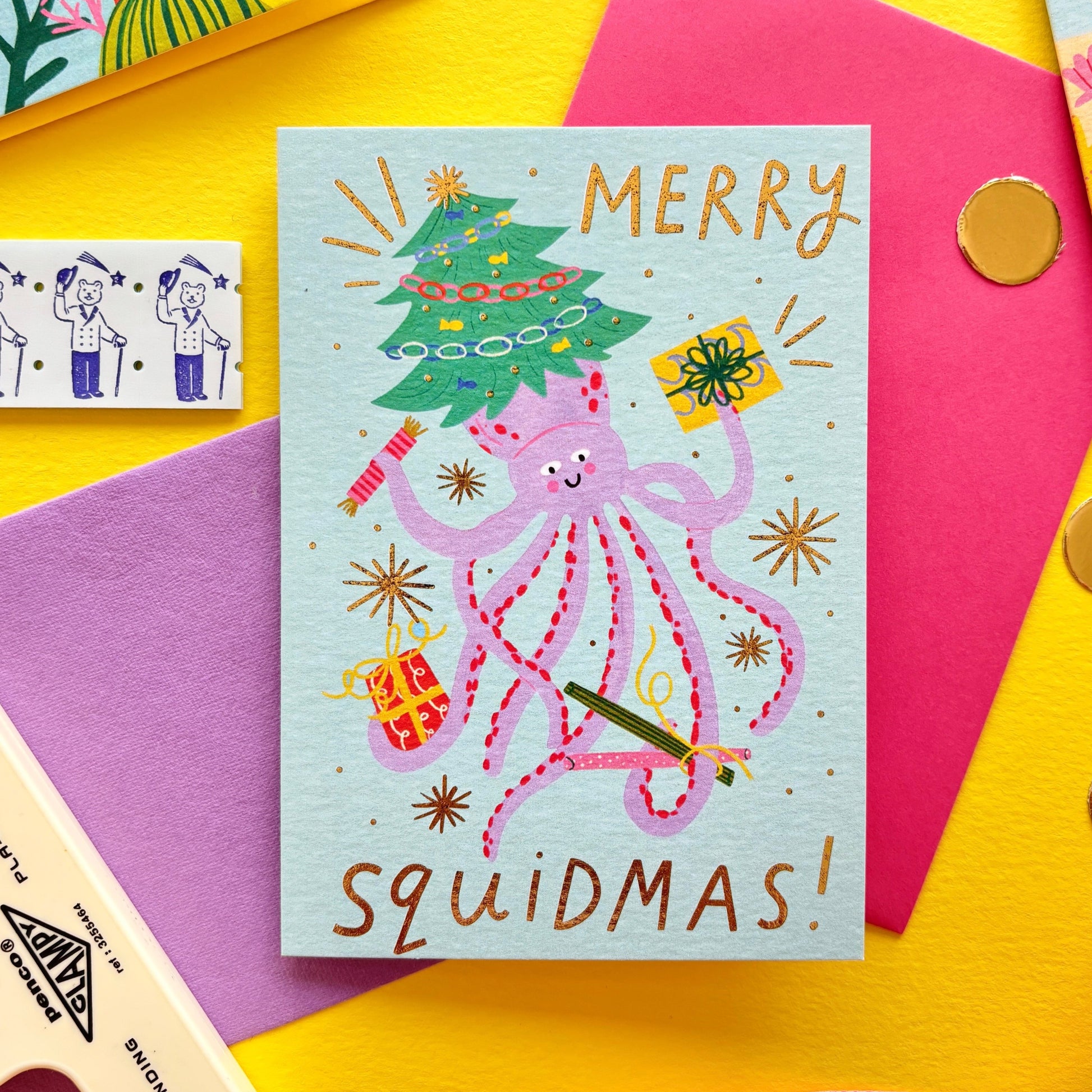 Funny Christmas card with pink squid holding presents and a Christmas tree – pun ‘Merry Squidmas!’ – festive ocean holiday card with gold foil stars on colourful flat lay background.