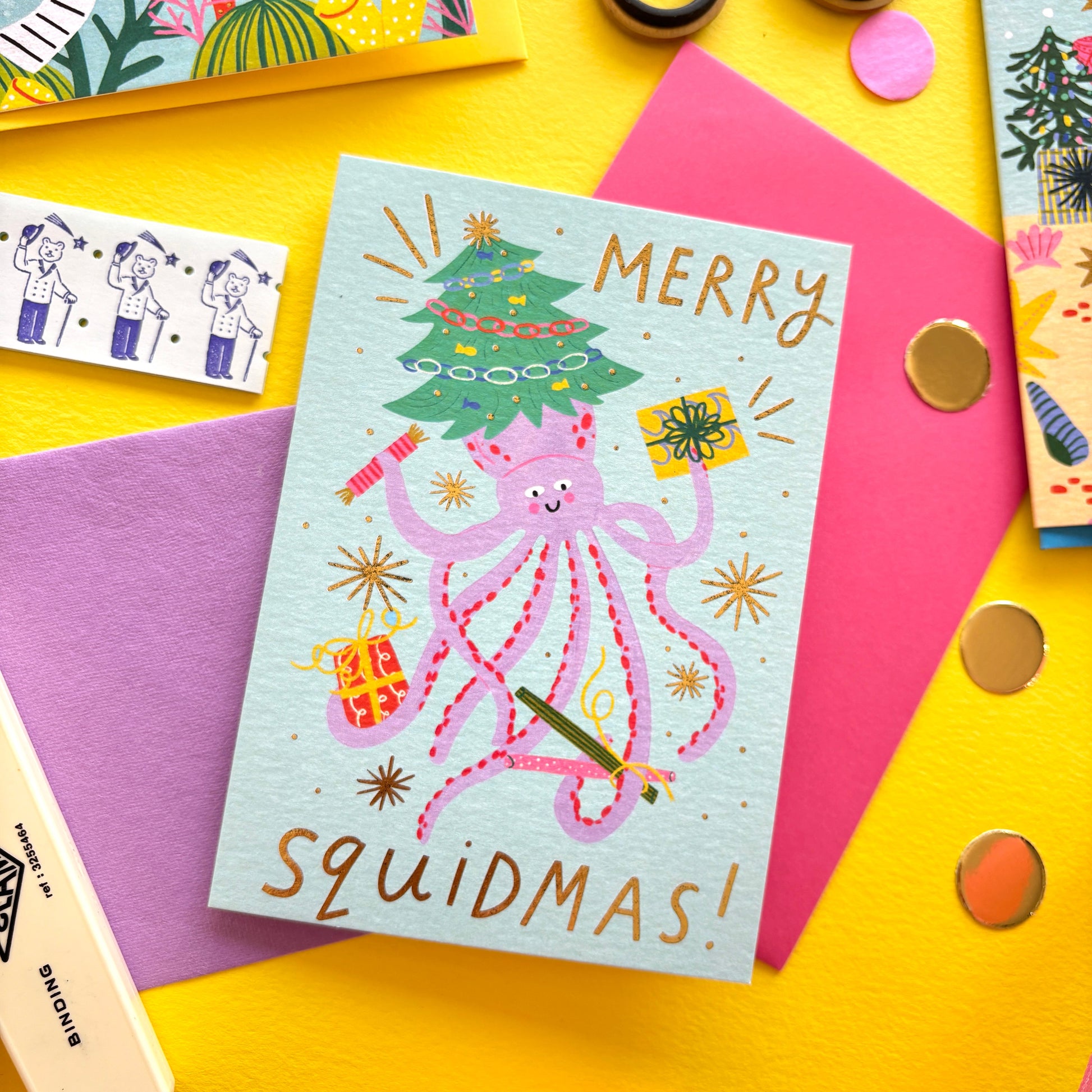 Funny Christmas card with pink squid holding presents and a Christmas tree – pun ‘Merry Squidmas!’ – festive ocean holiday card with gold foil stars on colourful flat lay background.
