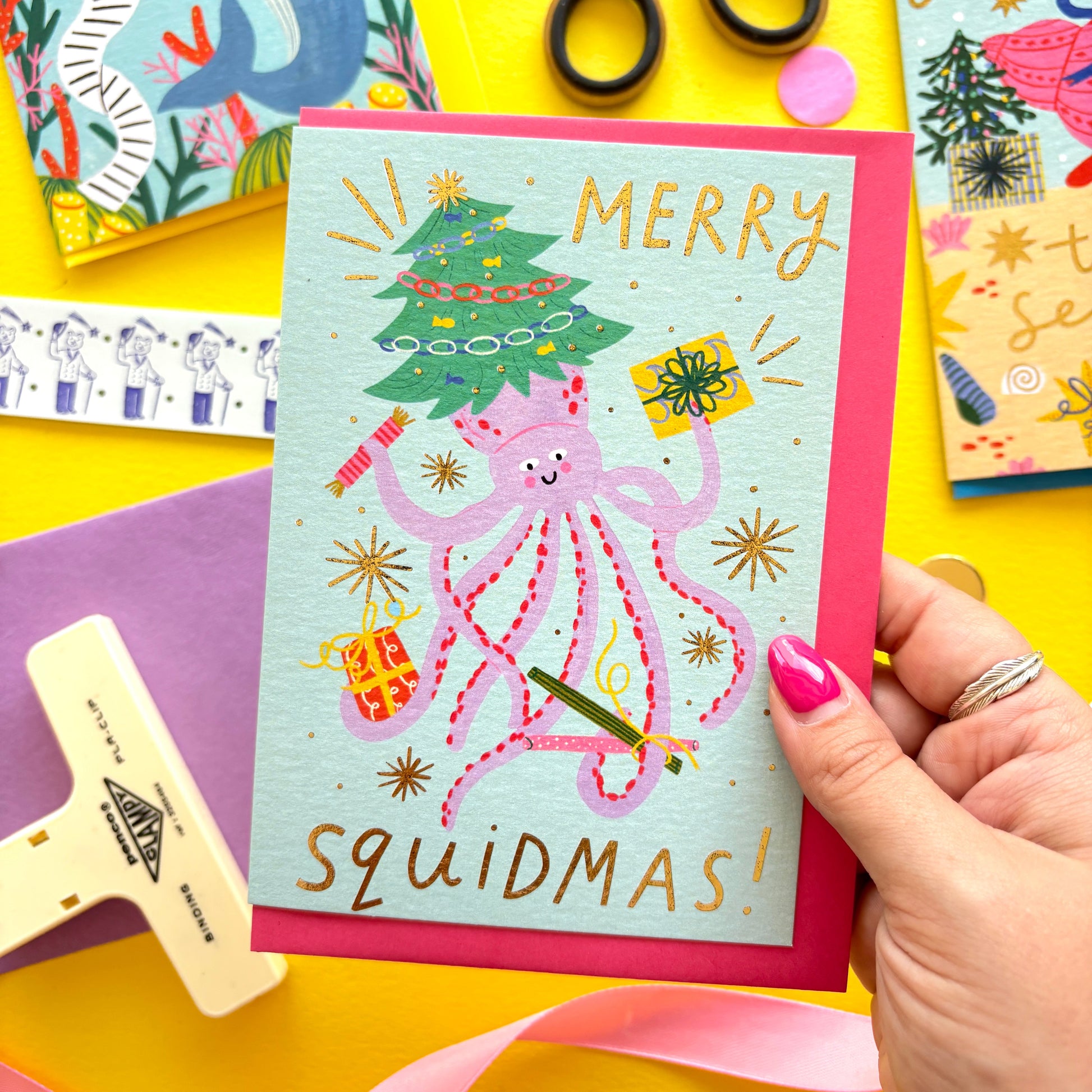 Funny Christmas card with pink squid holding presents and a Christmas tree – pun ‘Merry Squidmas!’ – festive ocean holiday card with gold foil stars on colourful flat lay background.