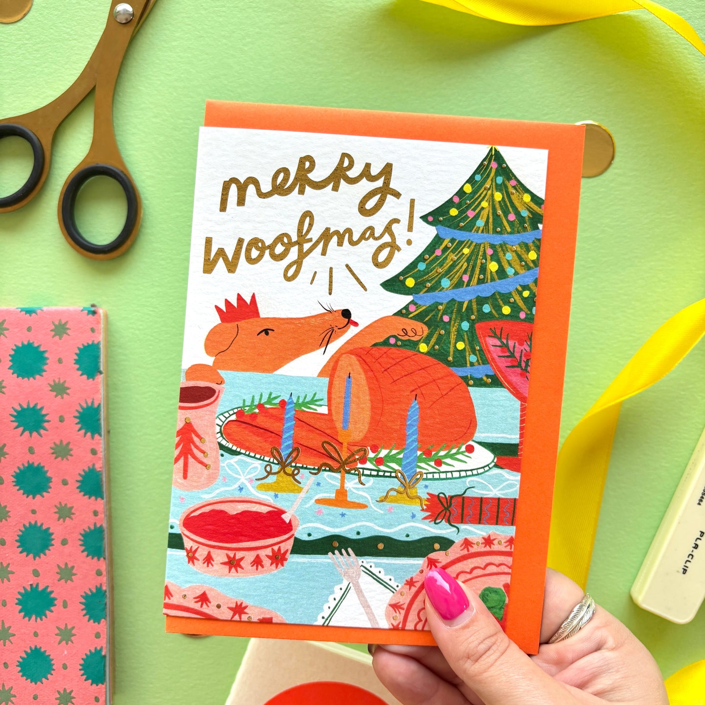 Merry Woofmas’ funny dog Christmas card featuring a cheeky dog licking its lips at a festive dinner table with roast turkey and gold foil details.