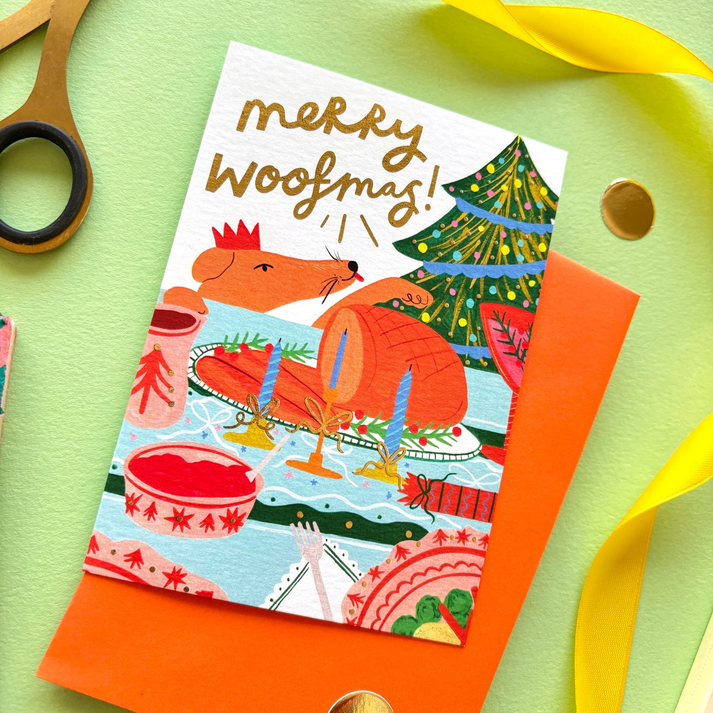Merry Woofmas’ funny dog Christmas card featuring a cheeky dog licking its lips at a festive dinner table with roast turkey and gold foil details.