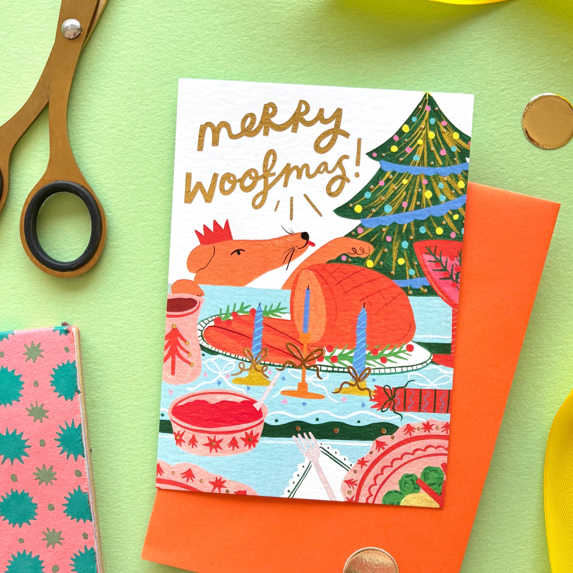 Merry Woofmas’ funny dog Christmas card featuring a cheeky dog licking its lips at a festive dinner table with roast turkey and gold foil details