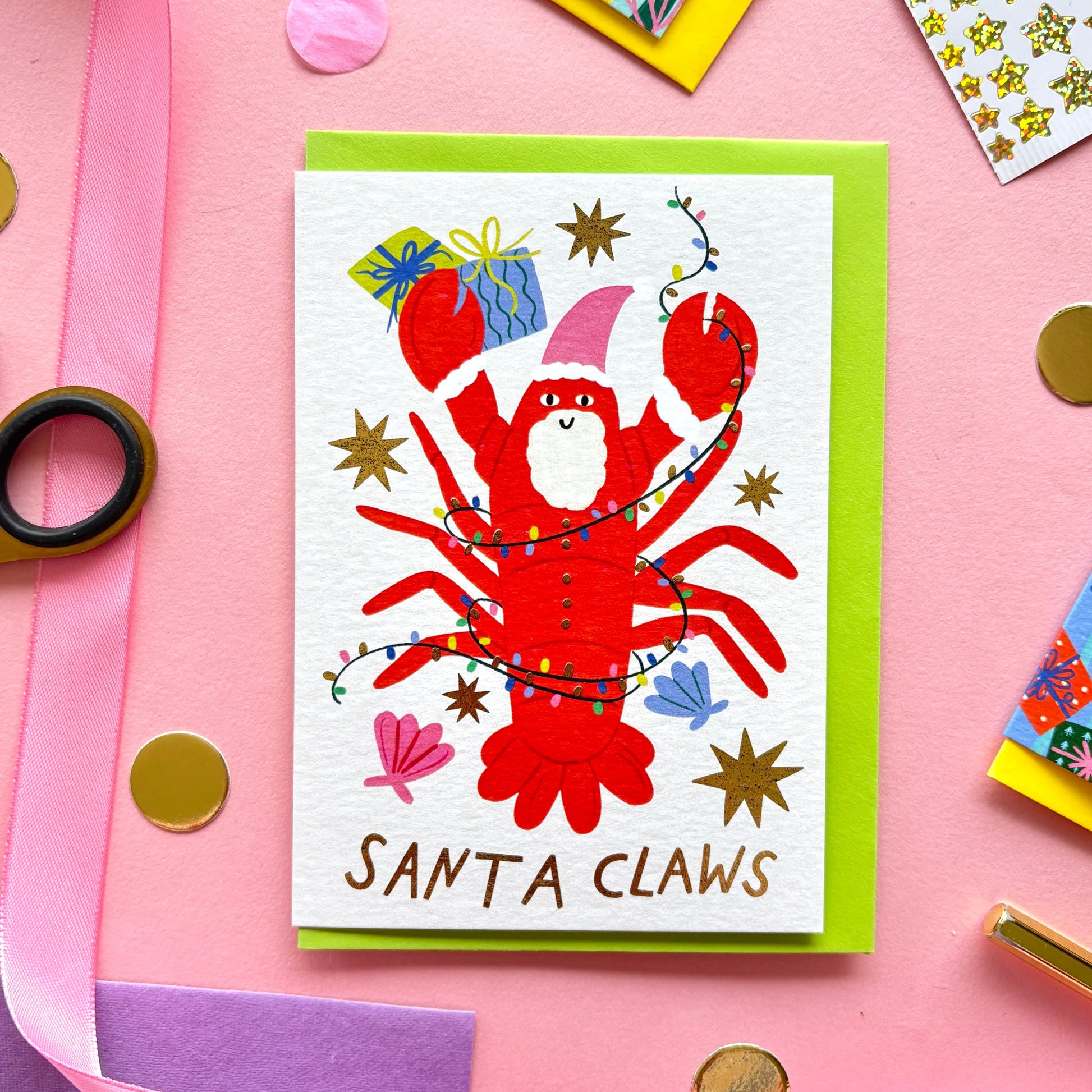 Playful flat lay photo of a funny Christmas card with the pun “Santa Claws!”. The design shows a smiling red lobster wearing a pink santa hat with a white beard. He is tangled in colourful lights and surrounded by shells, stars and colourful gifts. The card is paired with a bright pink envelope