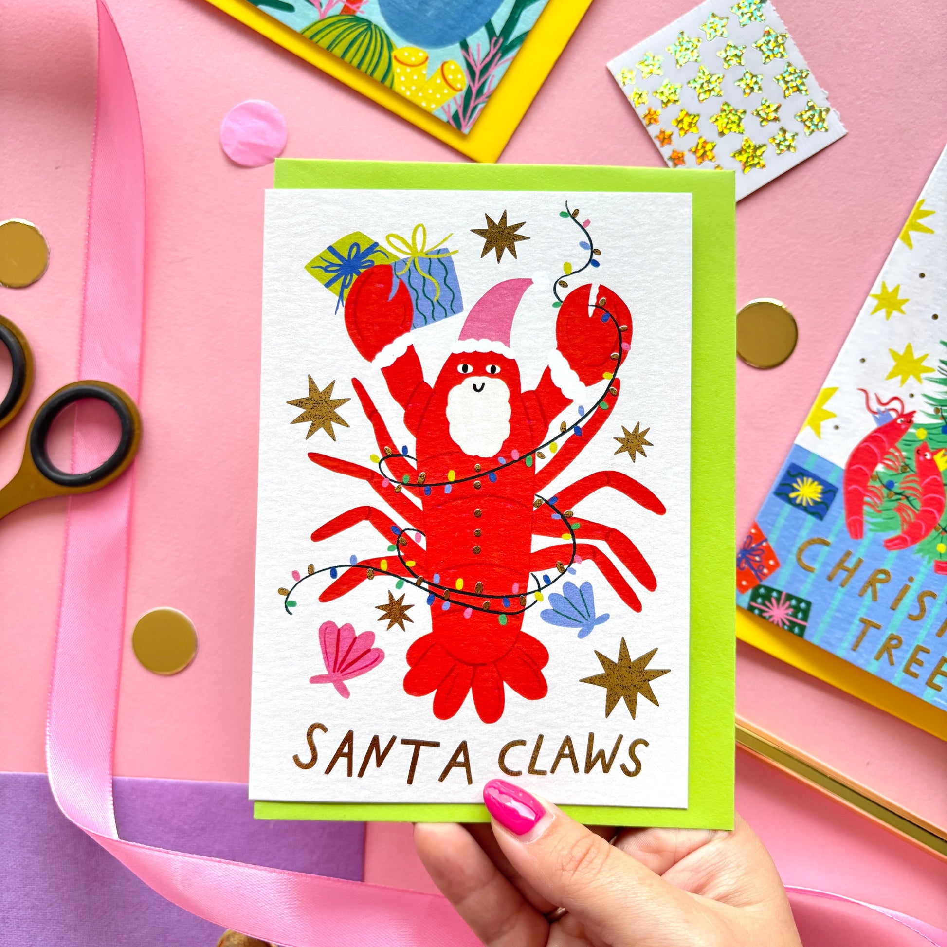 Playful flat lay photo of a funny Christmas card with the pun “Santa Claws!”. The design shows a smiling red lobster wearing a pink santa hat with a white beard. He is tangled in colourful lights and surrounded by shells, stars and colourful gifts. The card is paired with a bright pink envelope