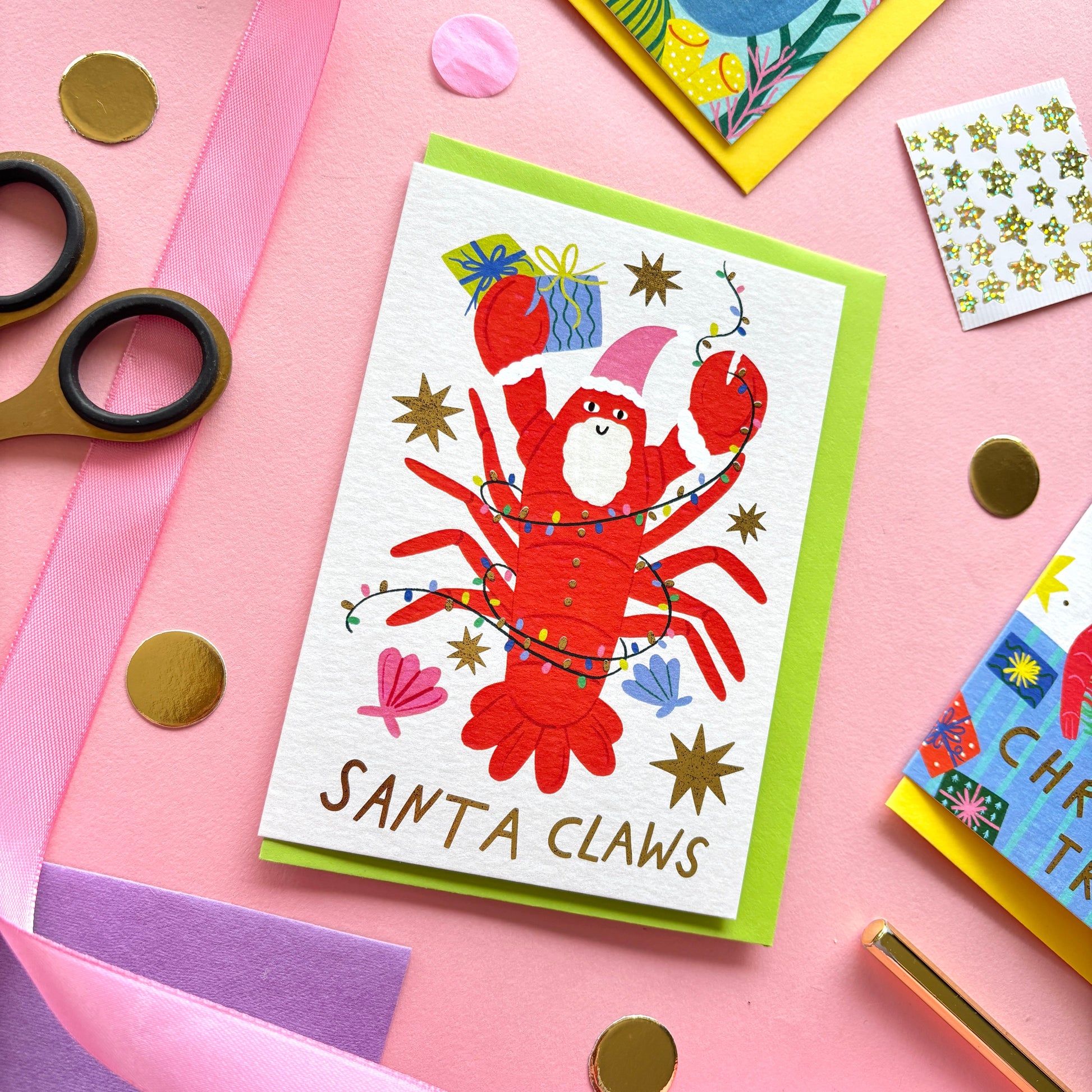 Playful flat lay photo of a funny Christmas card with the pun “Santa Claws!”. The design shows a smiling red lobster wearing a pink santa hat with a white beard. He is tangled in colourful lights and surrounded by shells, stars and colourful gifts. The card is paired with a bright pink envelope