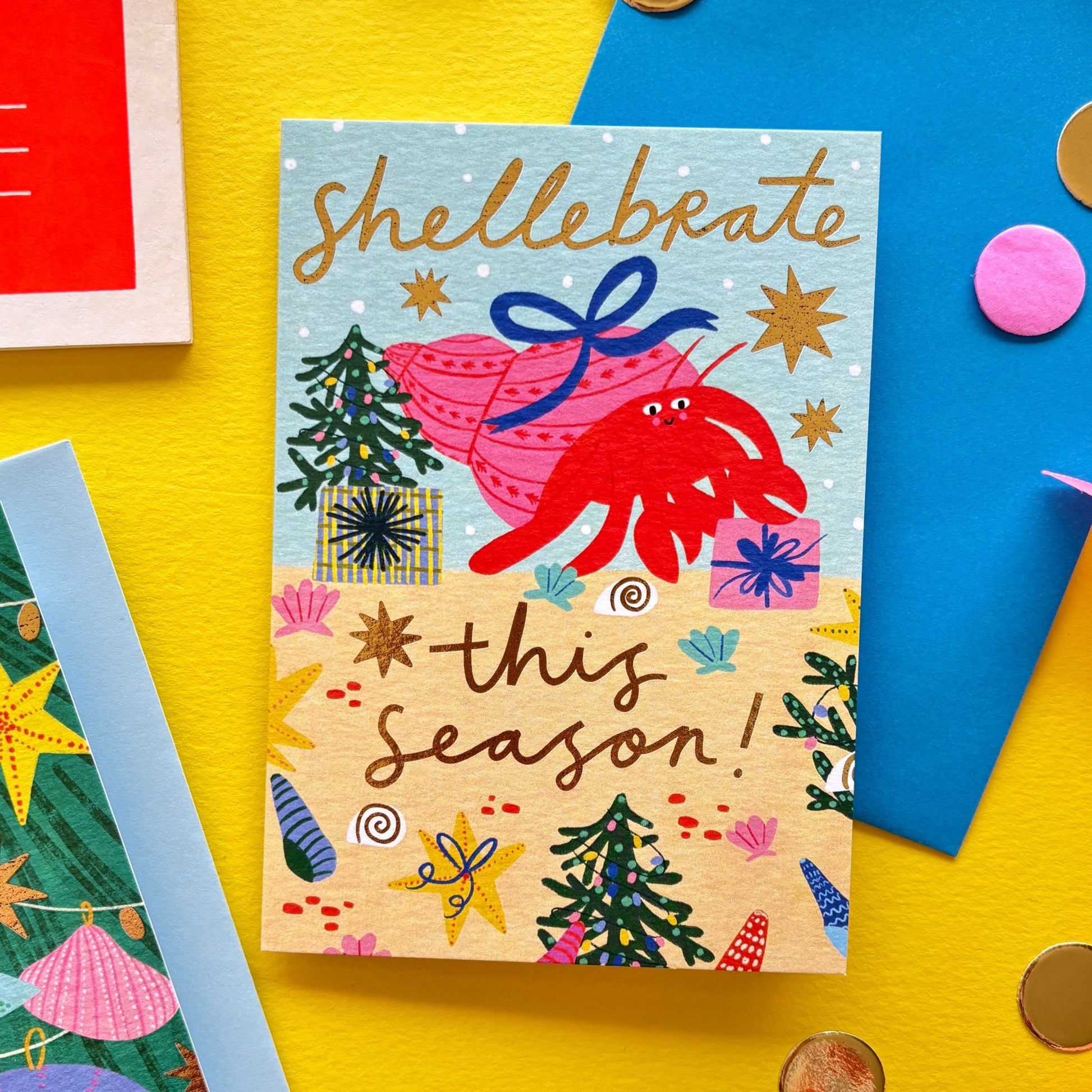 Funny Christmas card with red hermit crab holding a gift shell – pun ‘Shellebrate this season!’ – festive ocean holiday card with starfish, seashells, and Christmas trees on bright yellow background with ribbon, scissors, and envelopes