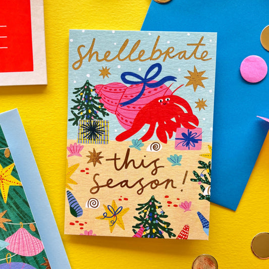 Funny Christmas card with red hermit crab holding a gift shell – pun ‘Shellebrate this season!’ – festive ocean holiday card with starfish, seashells, and Christmas trees on bright yellow background with ribbon, scissors, and envelopes