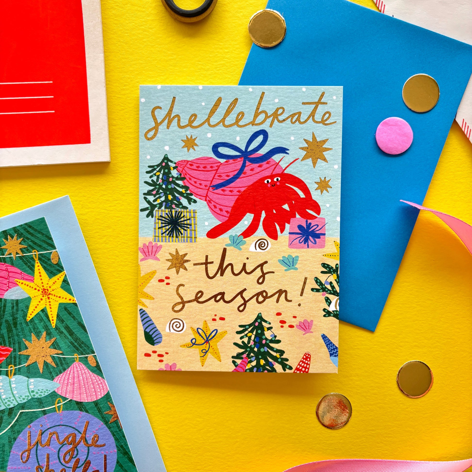 Funny Christmas card with red hermit crab holding a gift shell – pun ‘Shellebrate this season!’ – festive ocean holiday card with starfish, seashells, and Christmas trees on bright yellow background with ribbon, scissors, and envelopes