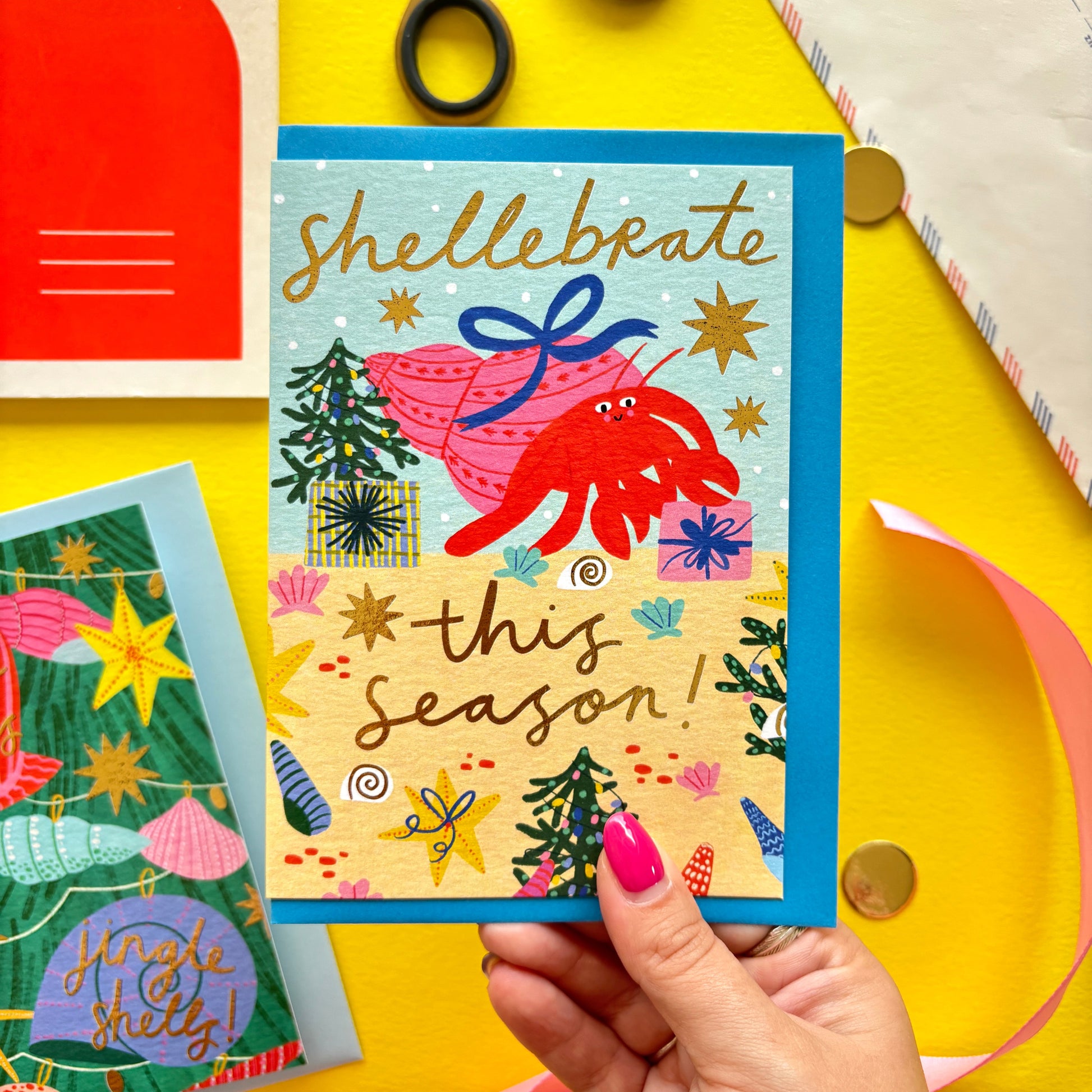 Funny Christmas card with red hermit crab holding a gift shell – pun ‘Shellebrate this season!’ – festive ocean holiday card with starfish, seashells, and Christmas trees on bright yellow background with ribbon, scissors, and envelopes