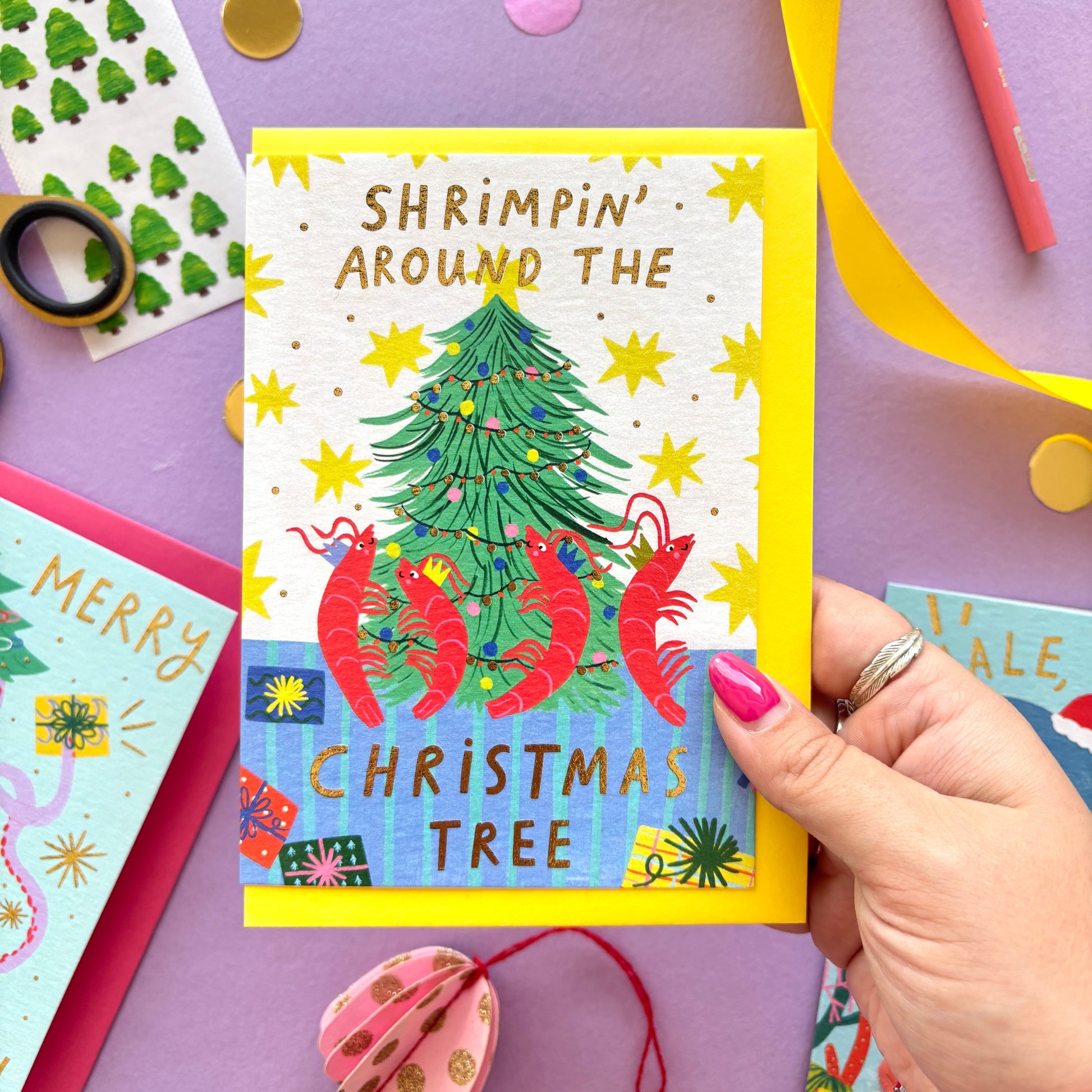 Funny Christmas card with red shrimp dancing around a Christmas tree – pun ‘Shrimpin’ Around the Christmas Tree’ – festive ocean-themed holiday card with bright yellow border on pastel background.