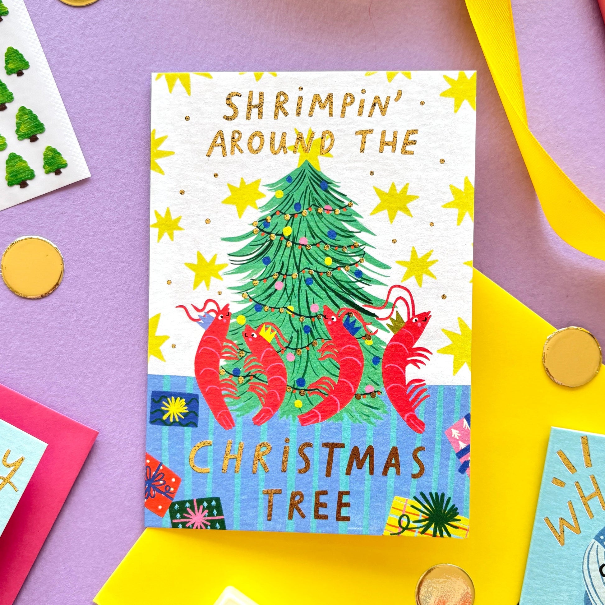 Funny Christmas card with red shrimp dancing around a Christmas tree – pun ‘Shrimpin’ Around the Christmas Tree’ – festive ocean-themed holiday card with bright yellow border on pastel background.