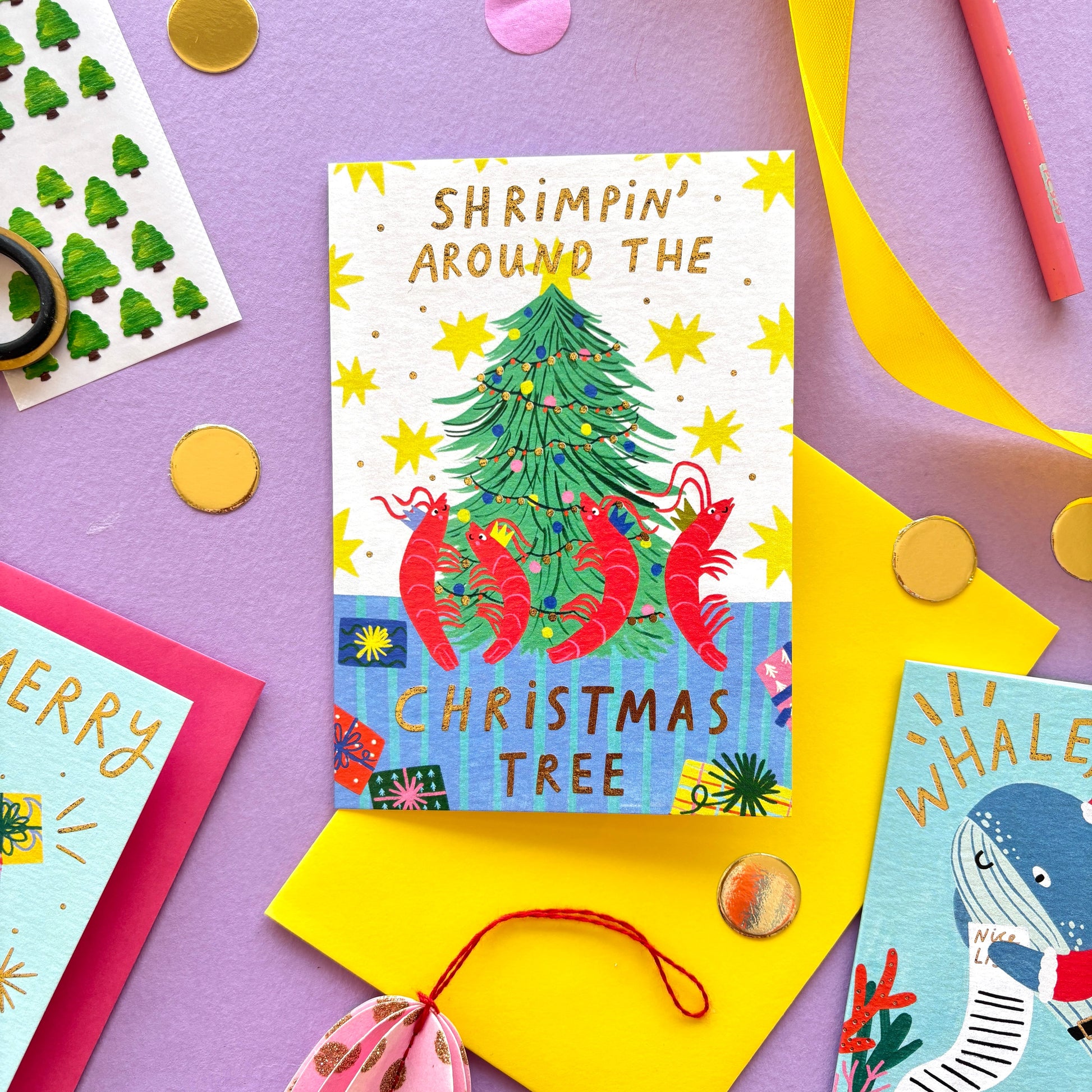 Funny Christmas card with red shrimp dancing around a Christmas tree – pun ‘Shrimpin’ Around the Christmas Tree’ – festive ocean-themed holiday card with bright yellow border on pastel background.