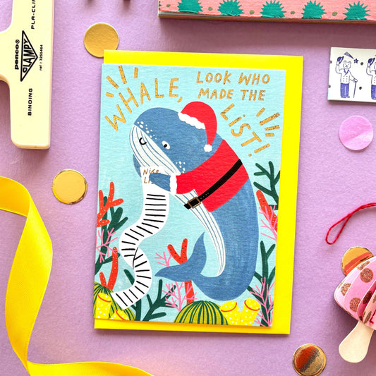 Funny Christmas card with a whale dressed as santa claus holding a long naughty and nice list surrounded by colourful coral along the reef- festive ocean-themed holiday card with bright blue background